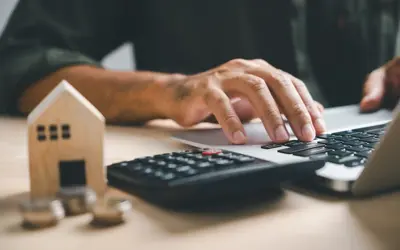 Person typing on a computer with a wooden house and coins stacked up in the foreground