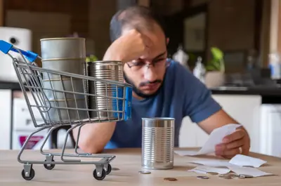 Man sitting in the kitchen holding receipt feeling frustrated due to inflation and increase of food costs with shopping cart in front of him holding food