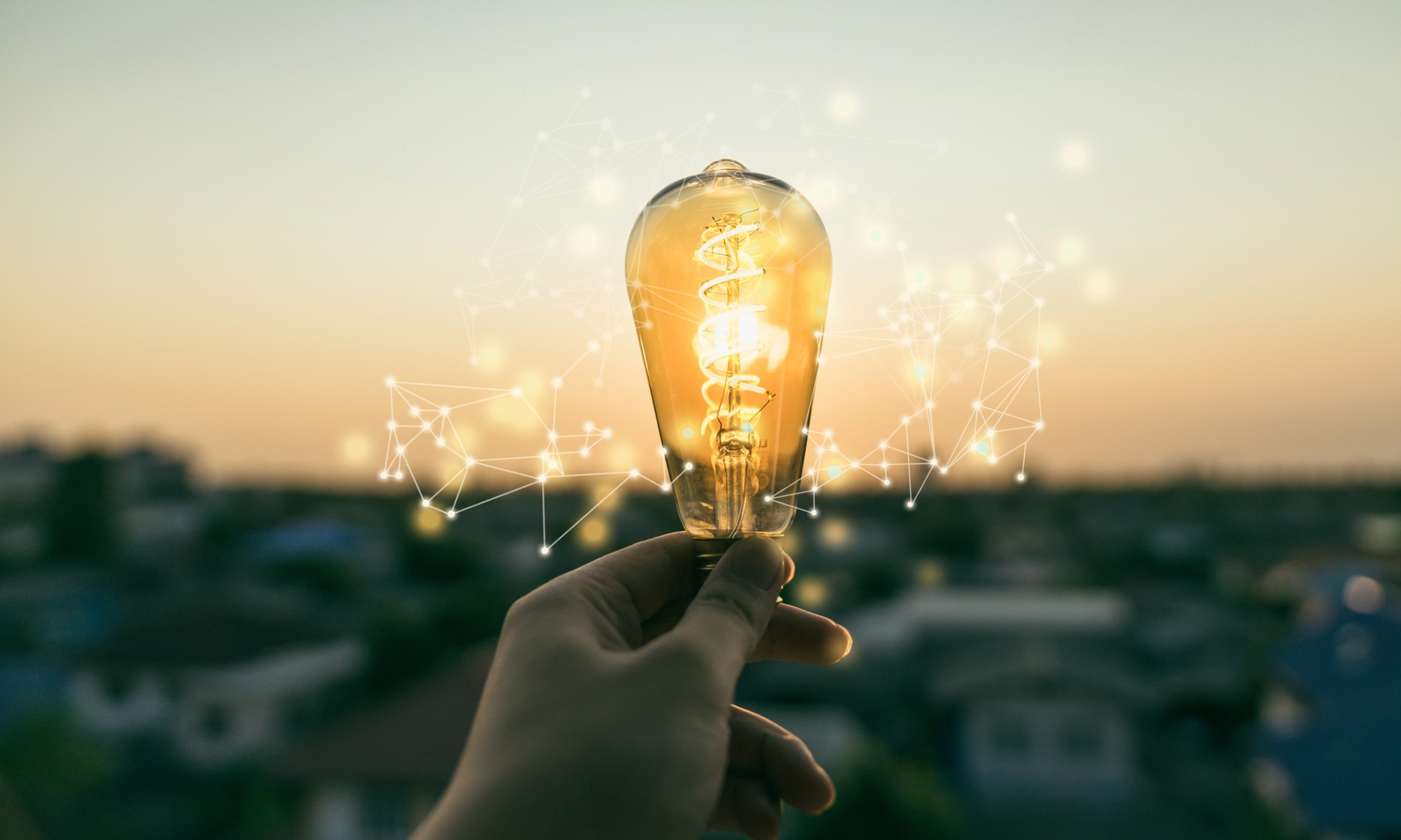 Close up of a hand holding a light bulb. There is a urban landscape in soft focus in the background.