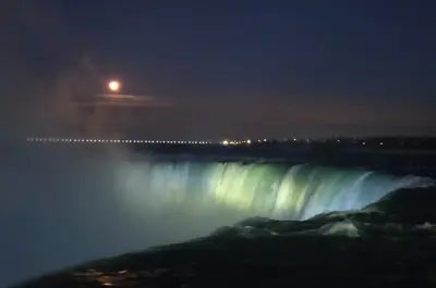 A full moon over Niagara Falls, illuminated by a line of lights in the distance