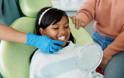 Young girl smiling and holding a mirror while being examined by a dentist.