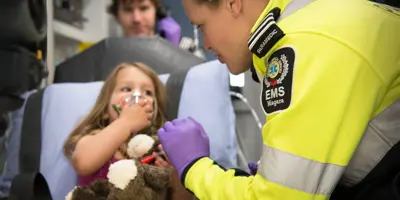 Paramedic tending to a child with a teddy bear and oxygen mask in an ambulance with another paramedic in the background