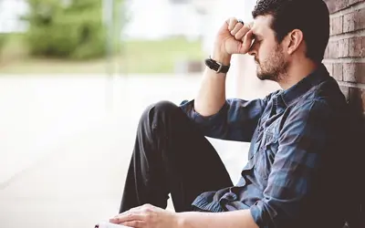 A depressed man sitting against a brick wall with his hand to his forehead.